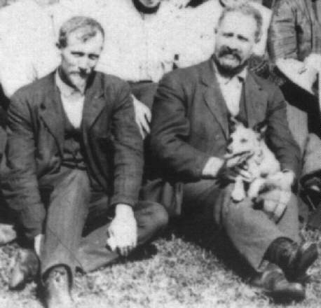two bearded men in dark suits sitting on the grass with the older man on the right holding a small dog