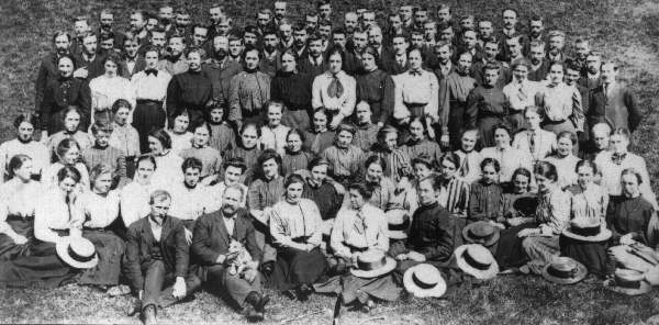 an old photograph showing a large group of men in jackets and women in shirtwaists and skirts with straw hats who are reclining on a grassy rise